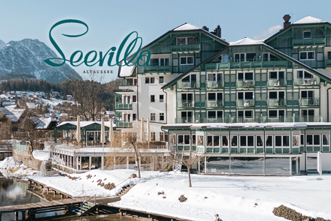 Ein großes Hotel am See mit grünen Balkonen liegt in einer verschneiten Landschaft mit Bergen im Hintergrund. In der Ecke erscheint der Text "Seevilla Altaussee".