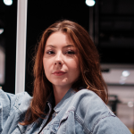 Eine Person mit langen braunen Haaren blickt in die Kamera. Sie trägt eine Jeansjacke. Der Hintergrund ist verschwommen, was auf eine Innenaufnahme schließen lässt.