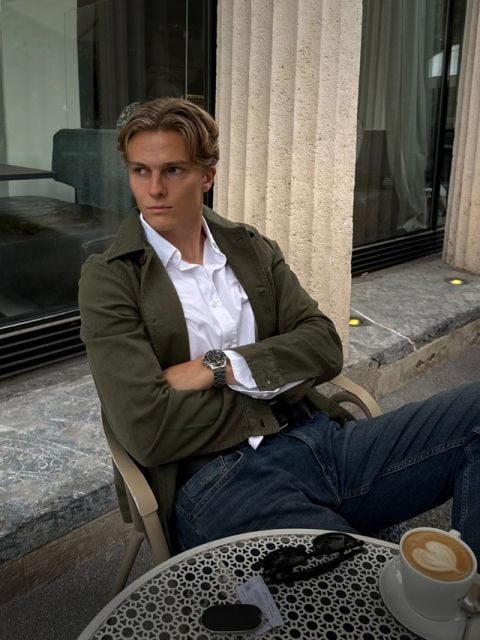Ein Mann in einem weißen Hemd und einer grünen Jacke sitzt an einem Tisch im Straßencafé, vor sich stehen Kaffee, eine Sonnenbrille und Schlüssel, und er schaut zur Seite.