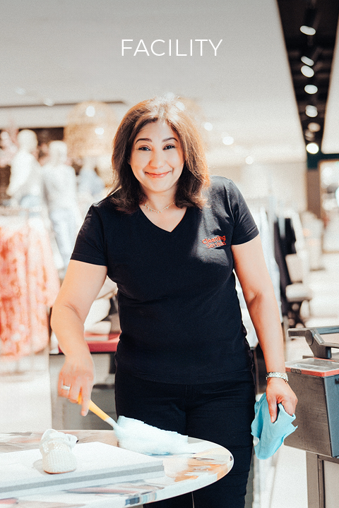 Eine Frau in einem schwarzen Hemd putzt lächelnd einen Tisch mit einem Staubtuch und einem Lappen in einem hellen, modernen Geschäft. Über ihr erscheint das Wort "FACILITY".