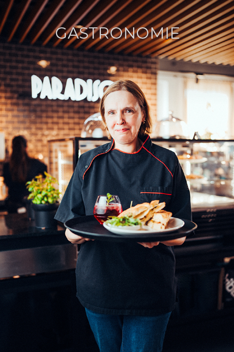 Ein Koch in schwarzer Uniform hält ein Tablett mit einem Gericht und einem Getränk in der Hand. Er steht in einem modernen Restaurant, in dem im Hintergrund die Schilder "PARADISO" und "GASTRONOMIE" zu sehen sind.
