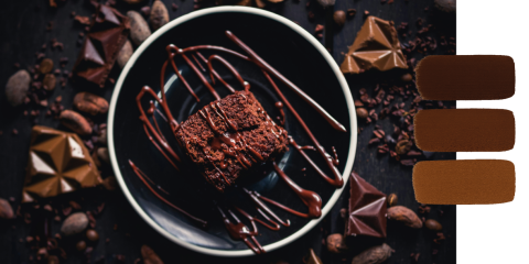 Ein Schokoladen-Brownie mit Schokoladensoße liegt auf einem schwarzen Teller, umgeben von Schokoladenstückchen und Kakaobohnen, mit drei braunen Farbflecken auf der rechten Seite des Bildes.