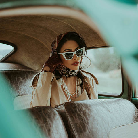 Eine elegante Frau mit Kopftuch, Sonnenbrille, Perlen und Trenchcoat sitzt auf dem Rücksitz eines Oldtimers und schaut mit selbstbewusstem Blick aus dem Fenster.