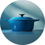 Ein runder, blau emaillierter gusseiserner Dutch Oven mit Deckel und Griffen steht auf einer passenden blauen Fläche, die von weichem, natürlichem Licht beleuchtet wird.
