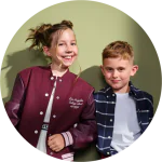 Zwei lächelnde Kinder stehen Seite an Seite vor einem grünen Hintergrund. Das Mädchen trägt eine kastanienbraune College-Jacke, der Junge ein marineblau kariertes Hemd über einem weißen T-Shirt. Sie sehen entspannt und fröhlich aus.