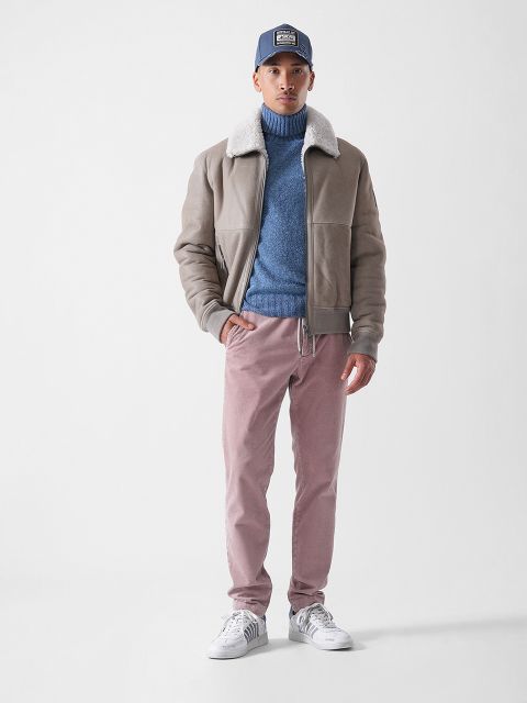 Ein Mann steht vor einem schlichten Hintergrund und trägt eine blaue Mütze, eine beigefarbene Shearling-Jacke, einen blauen Rollkragenpullover, eine hellrosa Hose und weiße Turnschuhe. Er blickt geradeaus, eine Hand in der Tasche.