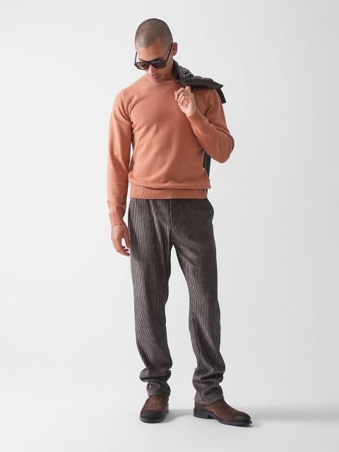 Ein Mann, der eine Sonnenbrille, einen pfirsichfarbenen Pullover, eine karierte Hose und braune Schuhe trägt, steht vor einem schlichten weißen Hintergrund und blickt mit einer über die Schulter gezogenen Jacke nach unten.