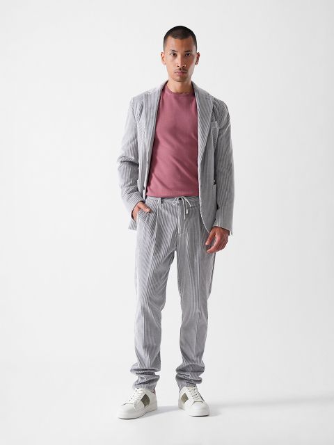 Ein Mann steht mit dem Gesicht nach vorne vor einem schlichten weißen Hintergrund. Er trägt einen hellgrau gestreiften Anzug mit einem lila T-Shirt und weißen Turnschuhen mit grauen Akzenten. Seine linke Hand steckt in der Hosentasche.