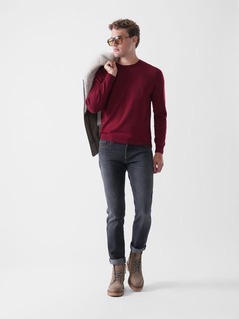 Ein Mann, der einen roten Pullover, dunkle Jeans, braune Stiefel und eine Sonnenbrille trägt, läuft mit einer braunen Jacke über der Schulter vor einem schlichten weißen Hintergrund.