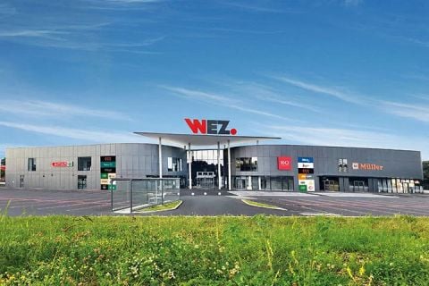 Ein modernes Einkaufszentrum mit einem großen "WEZ"-Schild auf dem Dach, einer grauen Fassade, verschiedenen Ladenschildern und einem klaren blauen Himmel darüber.