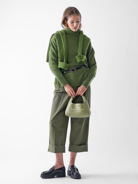 Eine Frau steht vor einem weißen Hintergrund und trägt einen grünen Rollkragenpullover, einen passenden grünen Pullover, der über ihre Schultern drapiert ist, eine olivgrüne Hose mit weitem Bein, schwarze Slipper und eine kleine blassgrüne Handtasche.