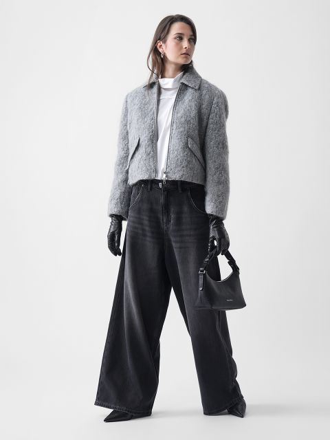 Eine Frau steht vor einem weißen Hintergrund und trägt eine kurz geschnittene graue Fuzzy-Jacke, schwarze Jeans mit weitem Bein, schwarze Handschuhe, schwarze spitze Schuhe und eine kleine schwarze Handtasche. Sie blickt mit einem neutralen Blick zur Seite.