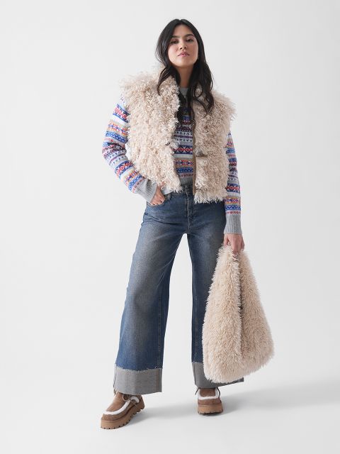 Eine Frau steht selbstbewusst in einer flauschigen beigen Weste, einem gemusterten Pullover, Jeans mit weitem Bein, hellbraunen Plateauschuhen und einer passenden flauschigen beigen Tasche vor einem schlichten weißen Hintergrund.