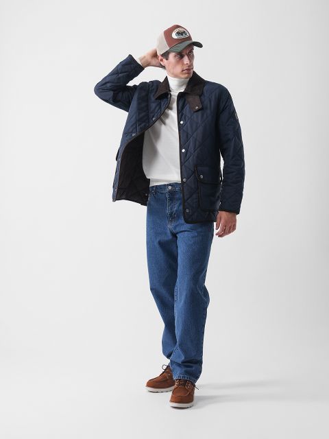 Ein Mann, der eine marineblaue Steppjacke, einen weißen Rollkragenpullover, blaue Jeans, braune Schuhe und eine beigefarbene Baseballmütze trägt, steht vor einem schlichten weißen Hintergrund und blickt nach links, wobei er mit einer Hand seine Mütze zurechtrückt.