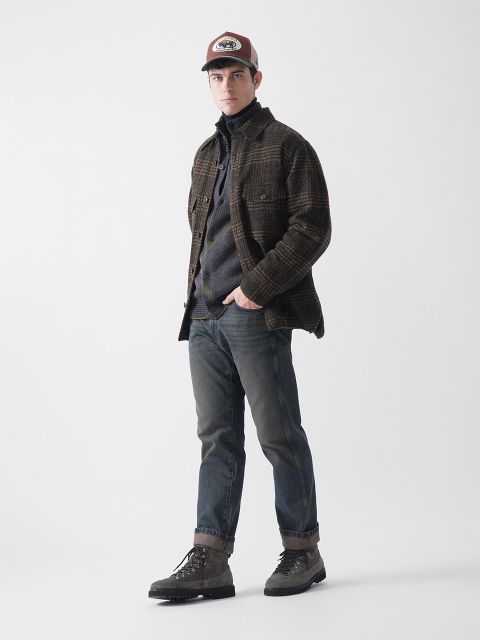 Ein Mann steht vor einem schlichten weißen Hintergrund und trägt eine karierte Jacke, einen dunklen Pullover, blaue Jeans, graue Stiefel und eine braune Baseballmütze. Er blickt mit neutraler Miene nach vorne, die Hände in den Taschen.