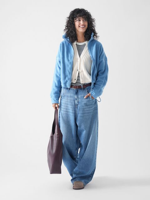 Eine Person mit lockigem Haar lächelt, während sie eine blaue Jacke mit Reißverschluss, ein gestreiftes Hemd, eine weiße Strickjacke, weite Bluejeans und braune Schuhe trägt. Sie trägt eine große dunkle Tragetasche und steht vor einem schlichten hellen Hintergrund.