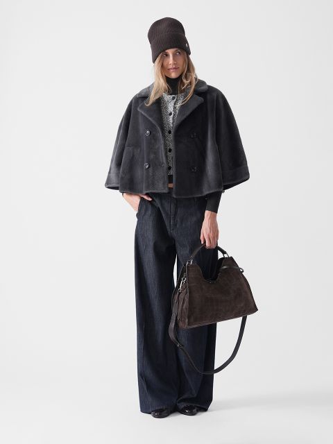 Eine Frau steht vor einem weißen Hintergrund, trägt eine dunkelbraune Mütze, einen kurzen grauen Pelzmantel, dunkle Jeans mit weitem Bein und schwarze Schuhe und hält eine große dunkelbraune Handtasche.