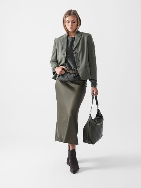 Eine Frau, die einen olivgrünen Blazer, ein dunkles Oberteil, einen olivgrünen Midirock und Stiefeletten trägt, hält eine große grüne Handtasche und steht vor einem schlichten weißen Hintergrund.