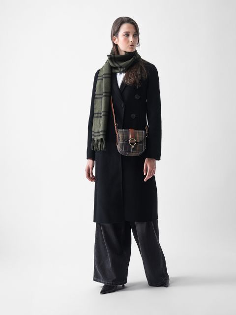 Eine Frau steht vor einem schlichten Hintergrund und trägt einen langen schwarzen Mantel, eine schwarze Hose mit weitem Bein, schwarze spitze Schuhe, einen grün karierten Schal und eine kleine karierte Tasche. Sie blickt leicht nach links.