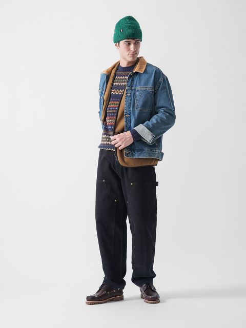 Eine Person, die eine grüne Mütze, einen gemusterten Pullover, eine Jeansjacke mit braunem Kragen, eine weite schwarze Hose und braune Schuhe trägt, steht vor einem schlichten weißen Hintergrund.