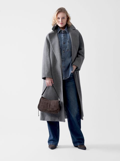 Eine Frau steht vor einem weißen Hintergrund, trägt einen langen grauen Mantel über einem Jeanshemd und Jeans mit weitem Bein und hält eine braune Handtasche in der Hand. Sie hat blondes Haar und schaut mit einem leichten Lächeln in die Kamera.
