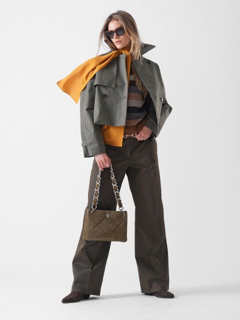 Eine Frau posiert selbstbewusst in einem olivgrünen Trenchcoat, einem senfgelben Schal, einem gestreiften Pullover, einer braunen Hose mit weitem Bein und einer Sonnenbrille und trägt eine gesteppte olivgrüne Handtasche mit einem klobigen Kettenriemen.