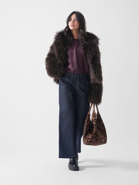 Eine Frau trägt eine zottelige braune Kunstpelzjacke, eine lila Bluse, dunkle Jeans mit weitem Bein und schwarze Schuhe, während sie vor einem schlichten weißen Hintergrund eine Tragetasche mit Leopardenmuster trägt.