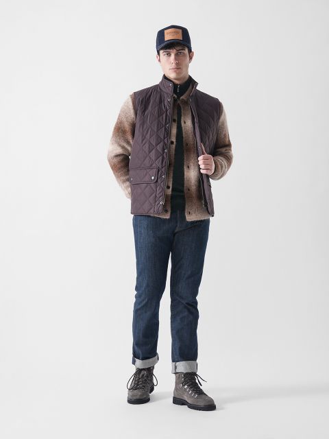 Ein Mann steht vor einem schlichten weißen Hintergrund und trägt eine dunkle Steppweste über einer braunen, wattierten Jacke, blaue Jeans mit Bündchen, graue Schnürstiefel und eine marineblaue Mütze mit einem braunen Aufnäher.