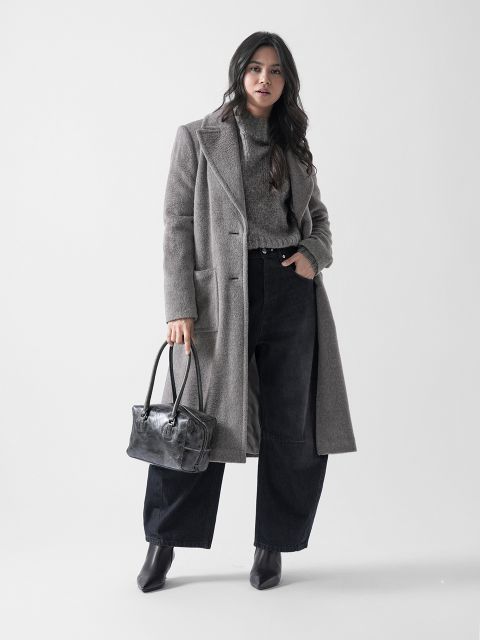 Eine Frau steht vor einem schlichten Hintergrund, trägt einen langen grauen Mantel, einen grauen Pullover, eine schwarze Hose mit weitem Bein und Stiefeletten und hält eine graue Handtasche an ihrer Seite.