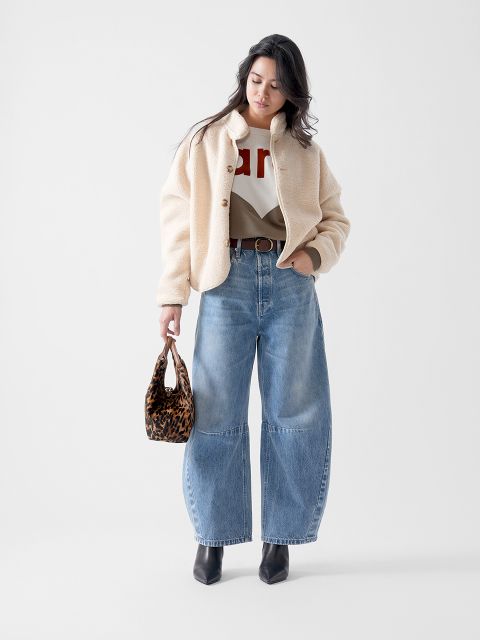 Eine Frau steht vor einem schlichten Hintergrund und trägt eine cremefarbene Sherpa-Jacke, weite Bluejeans, einen grafischen Pullover, schwarze Ankle Boots und eine kleine Handtasche mit Leopardenmuster. Sie blickt nach links.