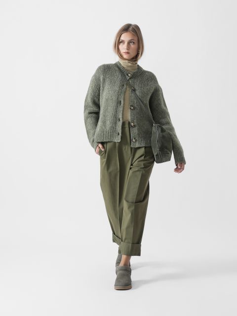 Eine Frau steht vor einem weißen Hintergrund und trägt eine grüne Strickjacke, einen olivgrünen Rollkragenpullover, eine weite olivgrüne Hose und graugrüne Schuhe, dazu eine grüne Clutch.