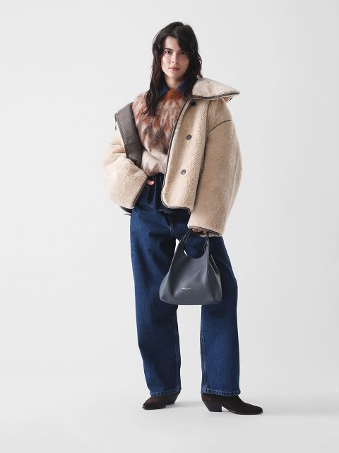 Eine Frau steht vor einem schlichten Hintergrund, trägt eine beigefarbene Shearling-Jacke, einen braunen Pelzschal, weite Bluejeans und dunkle hochhackige Stiefel und hält eine schwarze Handtasche in ihrer linken Hand.