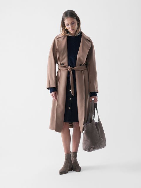 Eine Frau steht vor einem schlichten Hintergrund, trägt einen langen, kamelfarbenen Mantel über einem dunklen Kleid, braune Stiefeletten und hält eine taupefarbene Wildleder-Tasche. Sie blickt mit einem neutralen Blick nach unten.