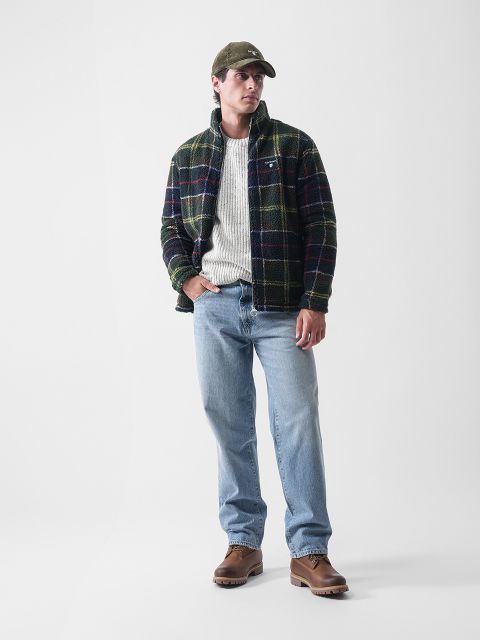 Ein Mann mit grüner Mütze, karierter Jacke, weißem Pullover, hellblauen Jeans und braunen Stiefeln steht vor einem schlichten weißen Hintergrund und blickt leicht zur Seite, eine Hand in der Tasche.