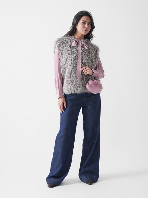 Eine Frau steht vor einem weißen Hintergrund und trägt eine rosafarbene Bluse mit Fliege, eine graue Kunstpelzweste, weit geschnittene Bluejeans, dunkle Schuhe und eine kleine rosafarbene Handtasche.