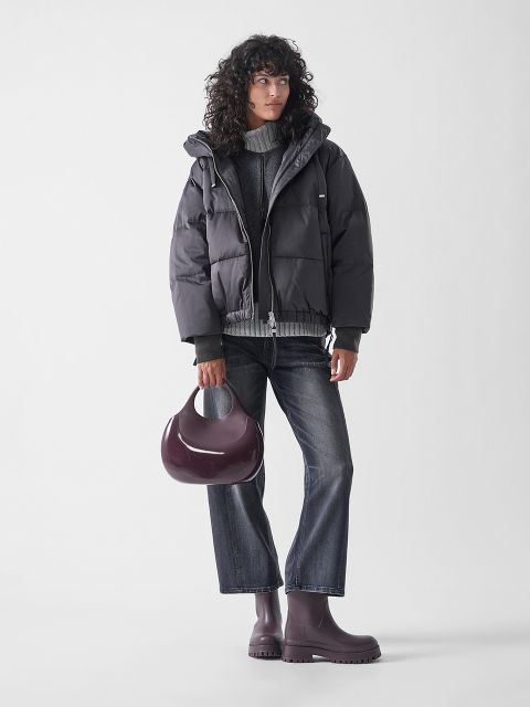 Eine Frau mit lockigem Haar trägt eine graue Pufferjacke, einen grauen Pullover, eine abgeschnittene dunkle Jeans und passende lila Regenstiefel. Sie hält eine glänzende, lila Handtasche und steht vor einem schlichten weißen Hintergrund.