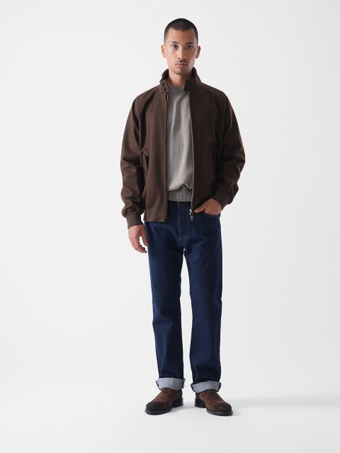 Eine Person steht vor einem schlichten weißen Hintergrund, trägt eine dunkelbraune Jacke, ein graues Hemd, dunkelblaue Jeans mit Manschetten und braune Schuhe und hat eine Hand in der Tasche.