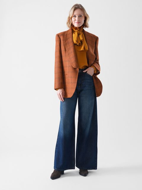 Eine Frau steht vor einem einfarbig weißen Hintergrund und trägt einen braun karierten Blazer, eine senfgelbe Bluse mit Schleife, eine weit geschnittene Bluejeans und braune Schuhe. Sie hat kurzes, gewelltes blondes Haar und ein entspanntes Lächeln.