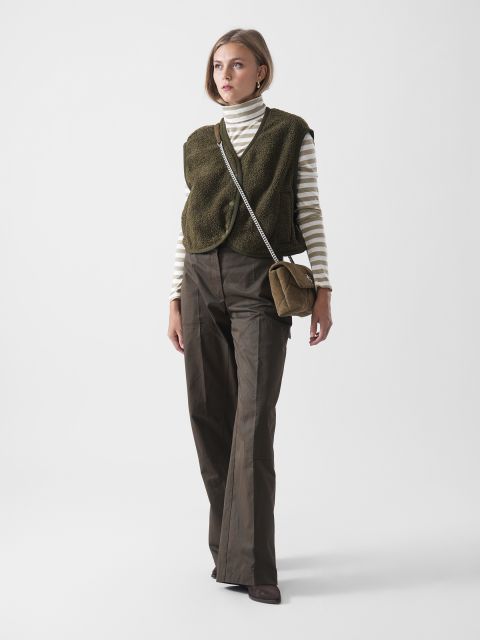 Eine Frau, die einen gestreiften Rollkragenpullover, eine olivgrüne strukturierte Weste, eine braune Hose mit weitem Bein und braune Schuhe trägt, steht mit einer braunen Umhängetasche vor einem schlichten weißen Hintergrund.