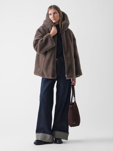 Eine Frau, die einen braunen Mantel mit Kapuze aus Kunstpelz, eine dunkle Jeans mit weitem Bein und Bündchen, schwarze Schuhe und eine braune Wildlederhandtasche trägt, steht vor einem schlichten weißen Hintergrund.