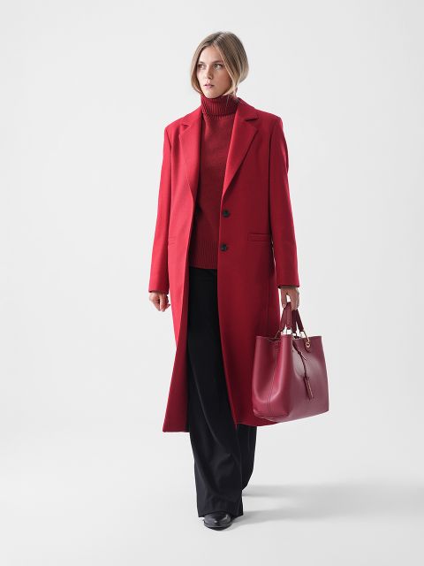Eine Frau steht vor einem schlichten Hintergrund, trägt einen langen roten Mantel über einem roten Rollkragenpullover, eine schwarze Hose und schwarze Schuhe und hält eine große rote Handtasche in der linken Hand.