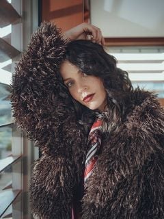 Eine Frau mit gewelltem dunklem Haar trägt einen weiten, flauschigen braunen Mantel und einen gemusterten Schal und lehnt sich an ein Fenster. Sie blickt mit erhobenem Arm in die Kamera, während das Sonnenlicht durch die Jalousien hinter ihr fällt.