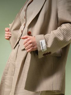 Eine Person, die einen beige-braun karierten Anzug mit einem weißen Etikett mit der Aufschrift "ABANDONABLE" an der Manschette trägt, hält das Revers der Jacke vor einem grünen Hintergrund.