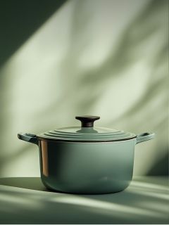 Ein grün emaillierter gusseiserner Dutch Oven mit Deckel und schwarzem Knauf steht auf einer Fläche, die von weichem natürlichem Licht mit unscharfen Schatten im Hintergrund beleuchtet wird.