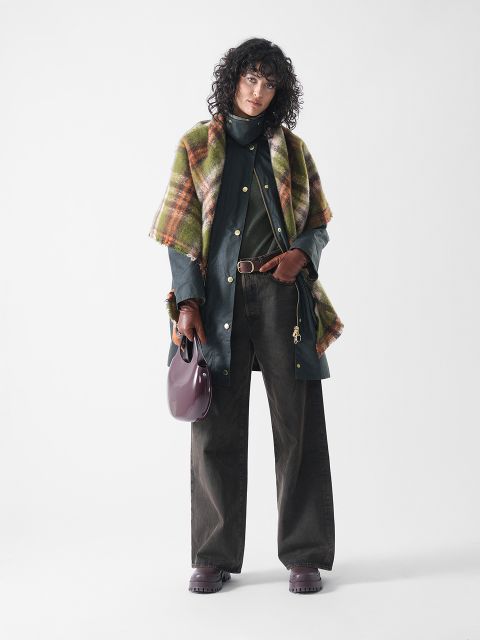 Eine Person mit lockigem Haar trägt einen grünen Mantel, eine weitbeinige braune Hose, braune Handschuhe, einen karierten Schal und dunkle Schuhe und hält eine glänzende bordeauxfarbene Handtasche in der Hand, während sie vor einem schlichten weißen Hintergrund steht.