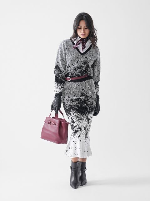 Eine Frau steht vor einem weißen Hintergrund und trägt einen schwarz-weiß gemusterten Pullover und Rock, schwarze Ankle Boots, Handschuhe, einen Gürtel, einen Schal und eine kastanienbraune Handtasche.