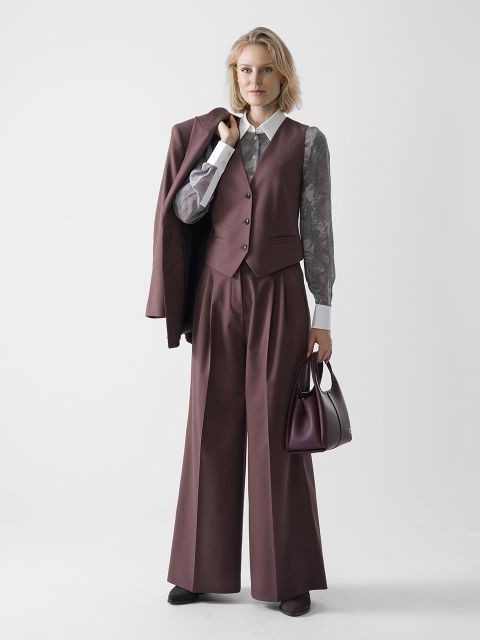 Eine Frau steht vor einem schlichten Hintergrund, trägt eine lila Weste und eine Hose mit weitem Bein, eine durchsichtige geblümte Bluse, hat eine passende Jacke über der Schulter und eine Handtasche in der anderen Hand.