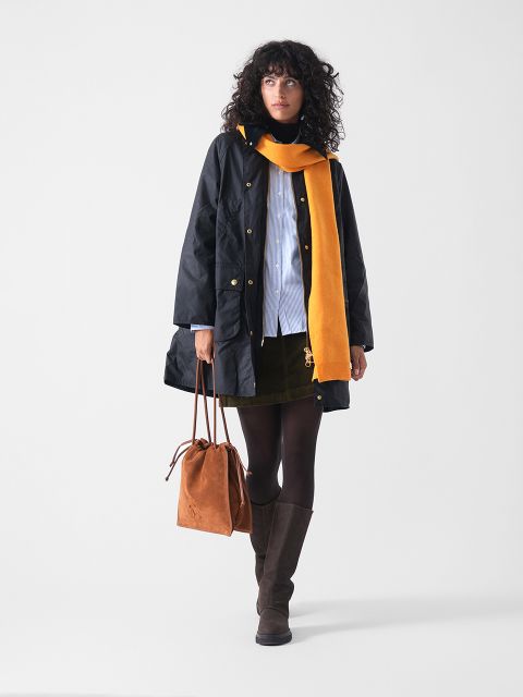 Eine Frau mit lockigem Haar trägt einen schwarzen Mantel, einen gelben Schal, ein hellblaues Hemd, einen dunklen Rock, eine braune Strumpfhose und kniehohe Stiefel. Sie trägt eine braune Wildlederhandtasche und steht vor einem schlichten weißen Hintergrund.