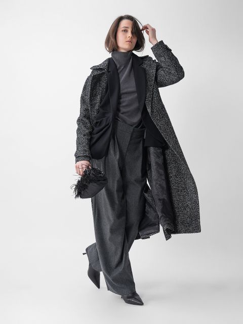 Eine Frau posiert vor einem schlichten Hintergrund. Sie trägt einen langen strukturierten Mantel über einem dunklen Rollkragenpullover und eine weit geschnittene Hose. Sie hält eine kleine, mit Federn besetzte Clutch und trägt spitze Absätze.