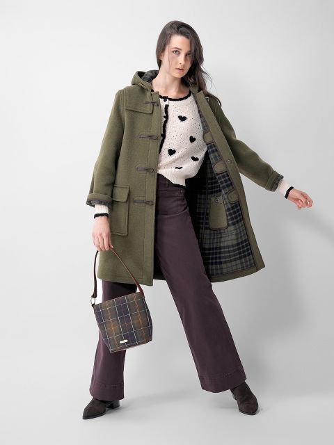 Eine Frau posiert in einem grünen Kapuzenmantel mit kariertem Futter, einem weißen Pullover mit schwarzen Herzen, einer braunen Hose mit weitem Bein und braunen Stiefeln und hält eine karierte Handtasche. Sie hat glattes braunes Haar und steht vor einem schlichten weißen Hintergrund.